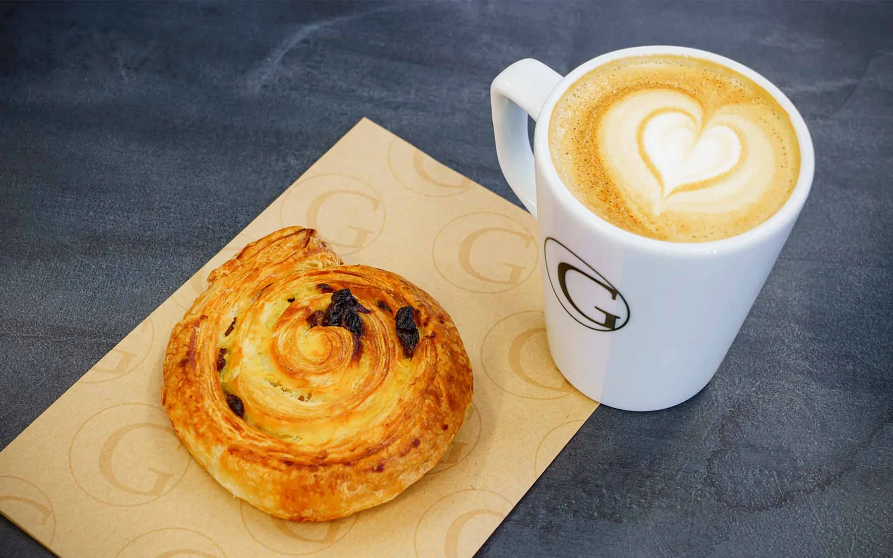 A latte in a white cup beside a swirled pastry on a paper bag