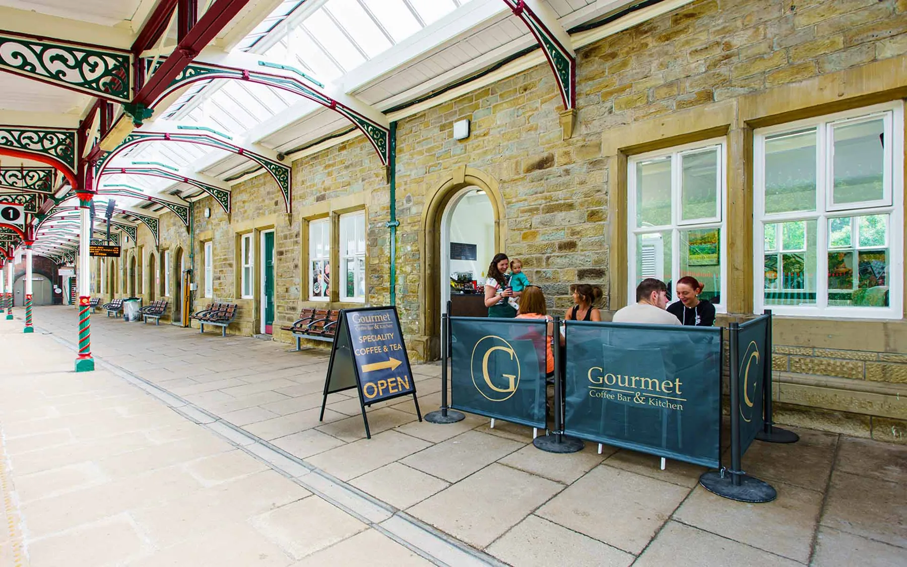A Gourmet Coffee Bar & Kitchen café on a station platform under the canopy, with an A-board sign outside