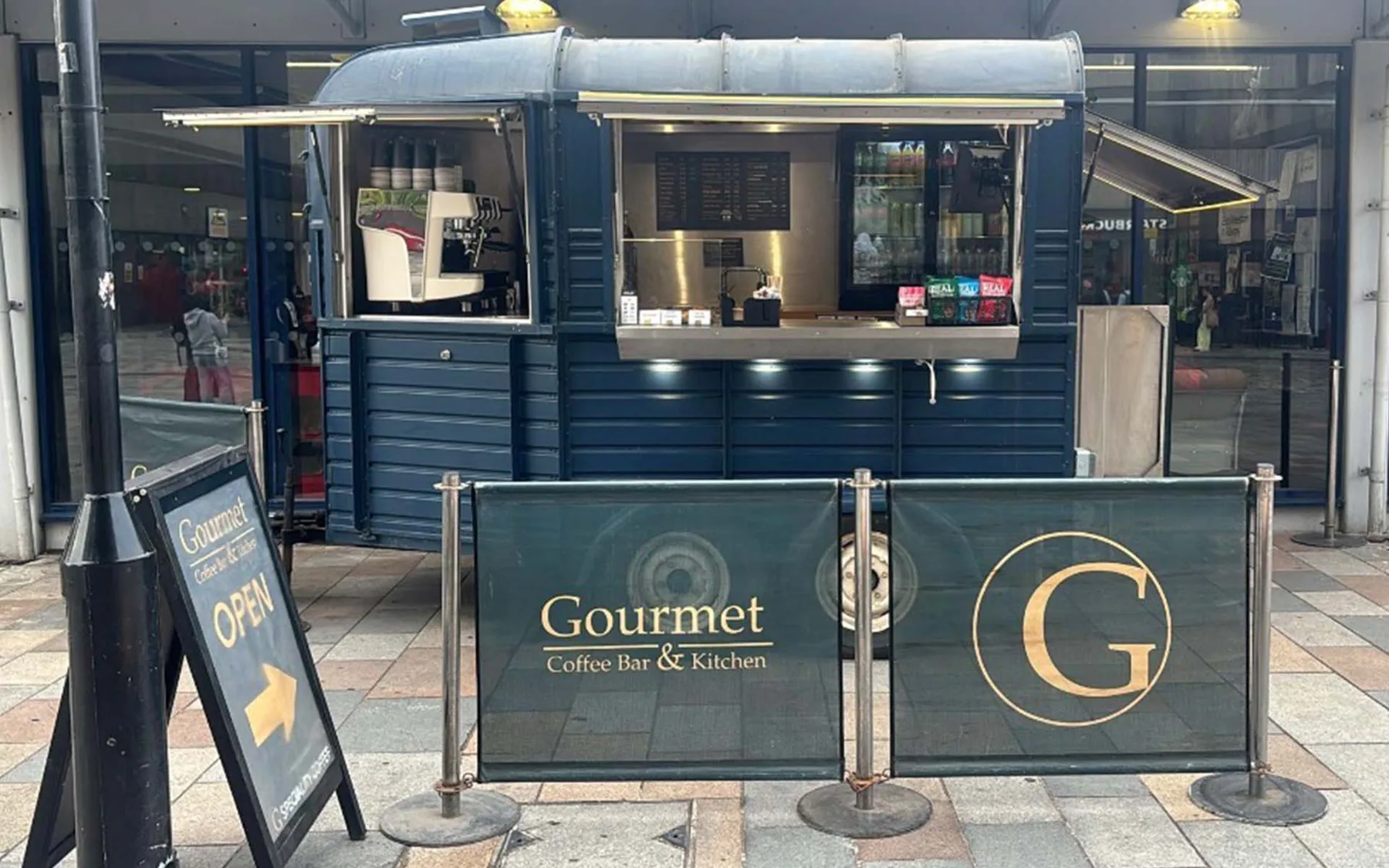 A navy Gourmet Coffee Bar & Kitchen kiosk with an A-board and cafe barriers